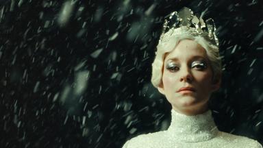 Porträtfoto einer weißblonden Frau mit silbrig glitzerndem Diadem in einem engen silbgrig glitzernden Rollkragen-Oberteil mit dickem silberfarbenem Lidschatten vor einem dunklen Hintergrund.