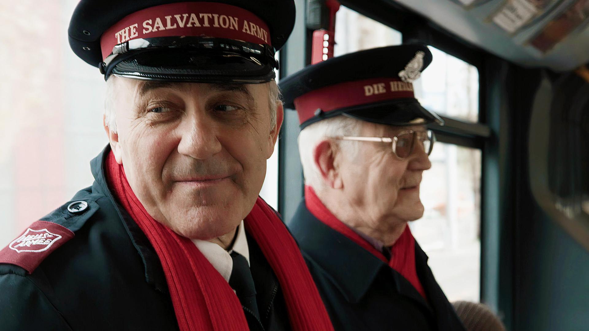 Zwei Mitglieder der Heilsarmee in Uniform mit roten Schals in einem öffentlichen Verkehrsmittel.