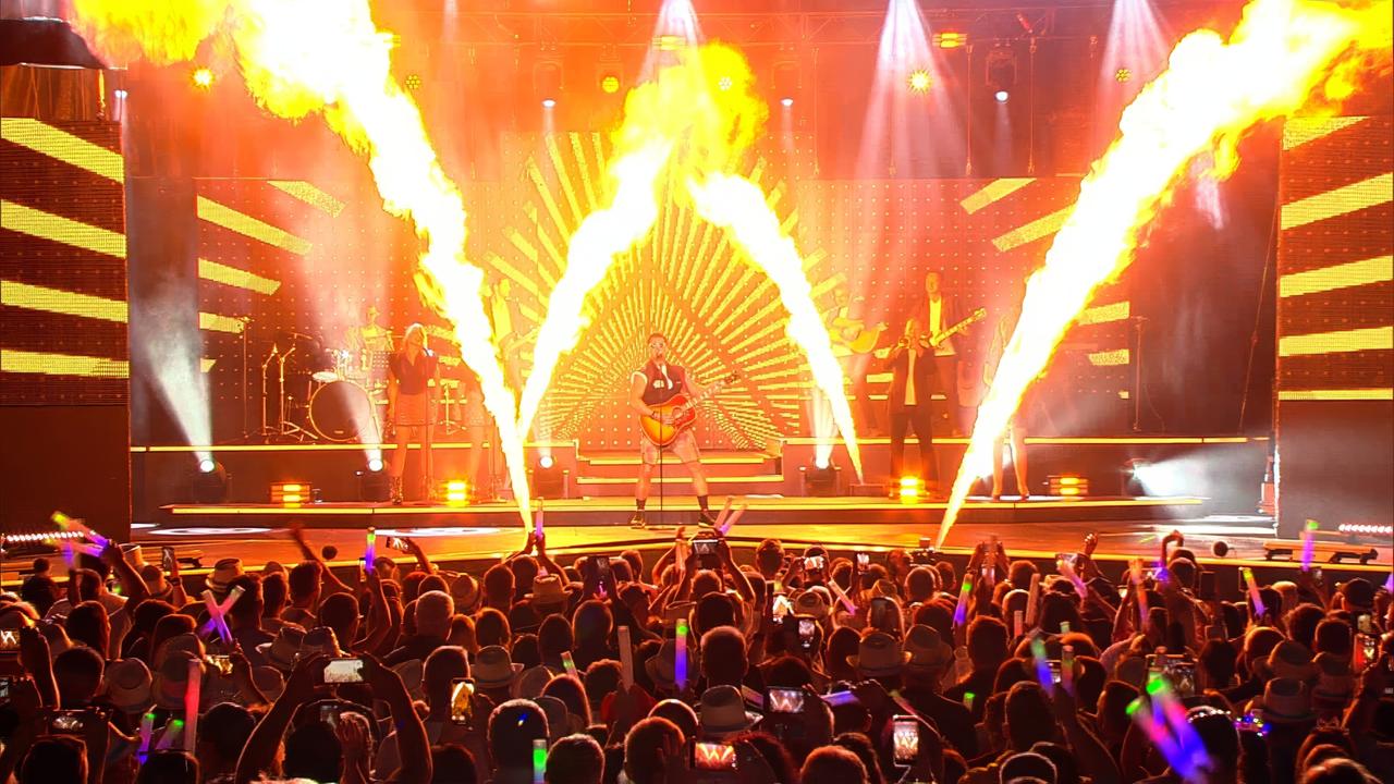 Das Bild zeigt eine Live-Performance auf einer Bühne, die mit bunten Lichtern und Effekten gestaltet ist. Zentrale Figur ist ein Musiker, der eine akustische Gitarre spielt und ein sportliches Outfit trägt. Um ihn herum stehen weitere Musiker, die in bunten Kostümen auftreten.   Im Hintergrund sind große LED-Wände sichtbar, die mit grafischen Mustern in warmen Farben leuchten. Auf der Bühne sind auch Pyrotechniken zu sehen, mit Flammen, die in die Luft schießen. Vor der Bühne steht eine große Menschenmenge, die begeistert mit Mobiltelefonen filmt und die Hände hebt, um die Energie der Aufführung zu unterstützen.   Das gesamte Setting vermittelt eine festliche und dynamische Atmosphäre, die typisch für Live-Konzerte im Musikgenre Schlager ist.