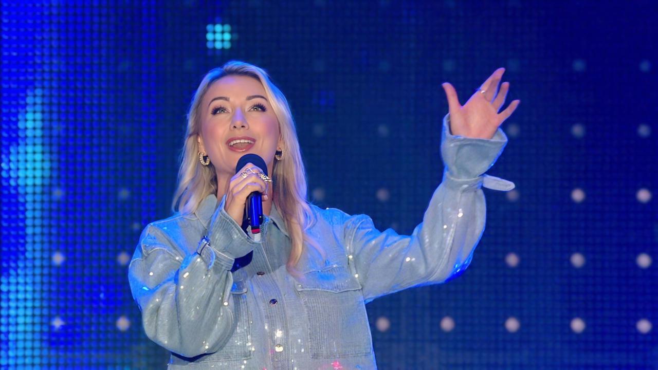 Das Bild zeigt eine singende Frau auf einer Bühne. Sie trägt eine glitzernde, hellblaue Jacke und hat lange, blonde Haare, die offen fallen. Die Frau hält ein Mikrofon in der linken Hand und hebt ihre rechte Hand in einer einladenden Geste. Im Hintergrund sind große, beleuchtete LED-Panels sichtbar, die mit Punkten und Lichtern in verschiedenen Farben dekoriert sind, was eine festliche Atmosphäre schafft. Das Bild vermittelt den Eindruck eines Livemusik-Events oder einer Bühnenaufführung.