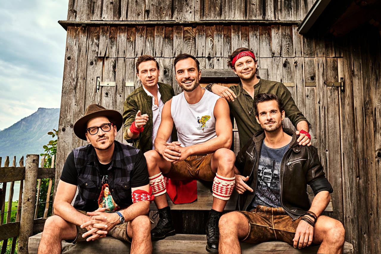 Das Bild zeigt fünf Männer, die vor einer rustikalen Holzstruktur posieren, die wahrscheinlich ein altes Gebäude oder eine Hütte ist. Die Holzverkleidung hat eine natürliche, verwitterte Optik.   Die Männer tragen verschiedene Kleidung, die zum ländlichen oder alpinen Stil passt. Einer von ihnen sitzt in der Mitte auf einer Holzbank und trägt ein weißes Tanktop sowie kurze Lederhosen. Zwei Männer stehen hinter ihm, einer trägt eine olivegrüne Jacke, während der andere ein Hemd mit langen Ärmeln in dunkler Farbe hat. Der dritte Mann auf der Bank hat ein Hemd mit kurzen Ärmeln und eine dreiviertellange Hose. Die Männer zeigen verschiedene Gesten, wie Daumen hoch oder Grinsen, und scheinen gut gelaunt zu sein.  Im Hintergrund ist eine grüne Landschaft zu sehen, die von sanften Hügeln und Bergen geprägt ist. Der Himmel ist bewölkt, was auf wechselhaftes Wetter hindeutet.   Das Bild vermittelt eine fröhliche Atmosphäre, die mit dem Lebensgefühl und der Kultur des Schlagers assoziiert werden kann.