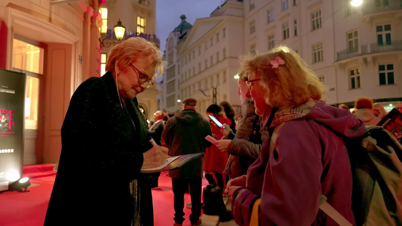 Das Bild zeigt eine Szene, die in der Dämmerung aufgenommen wurde, wahrscheinlich vor einem Veranstaltungsort. Im Vordergrund ist eine weibliche Person zu sehen, die Schrift auf einem Blatt Papier gibt. Diese Person hat kurze, blonde Haare und trägt eine schwarze, geflochtene Jacke. Um sie herum stehen mehrere Menschen, die auf ihre Smartphones schauen oder ebenfalls Autogramme erhalten möchten.  Im Hintergrund sind mehrstöckige Gebäude sichtbar, die in warmem Licht beleuchtet sind, was eine einladende Atmosphäre schafft. Einige Straßenlaternen sind ebenfalls eingeschaltet. Der Boden ist mit rotem Teppich ausgelegt, was auf eine besondere Veranstaltung hinweist.  Die gesamte Szene vermittelt eine lebhafte Stimmung mit Menschen, die auf Autogramme warten und die Interaktion mit einer bekannten Person genießen.