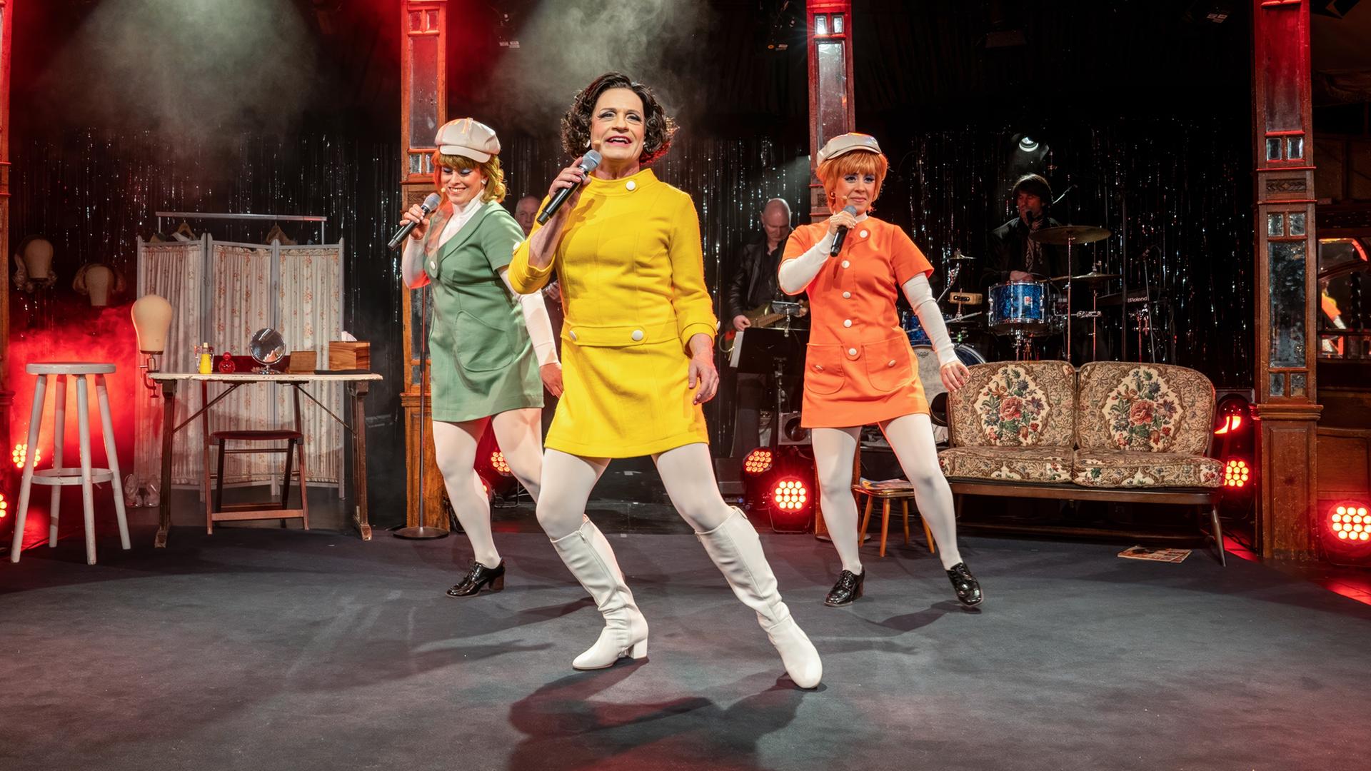 Auf dem Bild sind drei Frauen auf einer Bühne zu sehen, die eine Aufführung darbieten. Die Frauen tragen auffällige, farbenfrohe Kostüme: eine in einem leuchtend gelben Kleid, eine in grün und eine in orange, jeweils mit weißen Strumpfhosen und schwarzen Schuhen. Die Frau in Gelb steht im Vordergrund und hält ein Mikrofon, während die anderen beiden leicht im Hintergrund stehen und ebenfalls Mikrofone halten.   Im Hintergrund ist eine Bühne mit einer Lichtershow zu sehen, die mit roten und weißen Scheinwerfern beleuchtet ist. Der Bühnenboden ist schwarz und es gibt eine verhangene Kulisse aus glitzerndem Material hinter den Darstellerinnen. Eine einfache, retro angelehnte Couch steht seitlich auf der Bühne. An weiteren Bühnendetails erkennt man einige Musikinstrumente, und es gibt einen leicht nebligen Effekt in der Luft, der eine festliche Stimmung erzeugt.