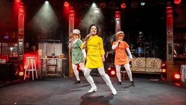 Auf dem Bild sind drei Frauen auf einer Bühne zu sehen, die eine Aufführung darbieten. Die Frauen tragen auffällige, farbenfrohe Kostüme: eine in einem leuchtend gelben Kleid, eine in grün und eine in orange, jeweils mit weißen Strumpfhosen und schwarzen Schuhen. Die Frau in Gelb steht im Vordergrund und hält ein Mikrofon, während die anderen beiden leicht im Hintergrund stehen und ebenfalls Mikrofone halten.   Im Hintergrund ist eine Bühne mit einer Lichtershow zu sehen, die mit roten und weißen Scheinwerfern beleuchtet ist. Der Bühnenboden ist schwarz und es gibt eine verhangene Kulisse aus glitzerndem Material hinter den Darstellerinnen. Eine einfache, retro angelehnte Couch steht seitlich auf der Bühne. An weiteren Bühnendetails erkennt man einige Musikinstrumente, und es gibt einen leicht nebligen Effekt in der Luft, der eine festliche Stimmung erzeugt.