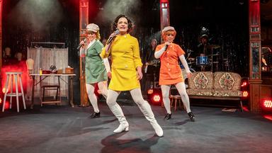 Auf dem Bild sind drei Frauen auf einer Bühne zu sehen, die eine Aufführung darbieten. Die Frauen tragen auffällige, farbenfrohe Kostüme: eine in einem leuchtend gelben Kleid, eine in grün und eine in orange, jeweils mit weißen Strumpfhosen und schwarzen Schuhen. Die Frau in Gelb steht im Vordergrund und hält ein Mikrofon, während die anderen beiden leicht im Hintergrund stehen und ebenfalls Mikrofone halten.   Im Hintergrund ist eine Bühne mit einer Lichtershow zu sehen, die mit roten und weißen Scheinwerfern beleuchtet ist. Der Bühnenboden ist schwarz und es gibt eine verhangene Kulisse aus glitzerndem Material hinter den Darstellerinnen. Eine einfache, retro angelehnte Couch steht seitlich auf der Bühne. An weiteren Bühnendetails erkennt man einige Musikinstrumente, und es gibt einen leicht nebligen Effekt in der Luft, der eine festliche Stimmung erzeugt.