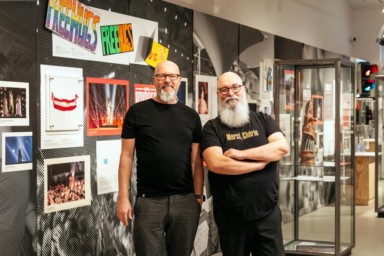 Das Bild zeigt zwei Männer, die in einem Raum stehen, der mit verschiedenen Ausstellungsstücken und Fotografien dekoriert ist. Sie befinden sich vor einer Wand, die mit einem grafischen Hintergrund in Schwarz und Weißen Punkten gestaltet ist. Darüber hängen bunte Schilder. Eines der auffälligsten Schilder zeigt die Aufschrift "FREE HUGS" in bunten Buchstaben.  Die beiden Männer tragen schwarze T-Shirts. Der Mann links hat eine Brille und einen kurzen, grauen Bart. Der Mann rechts hat ebenfalls eine Brille, einen weißen Bart und trägt ein T-Shirt mit der Aufschrift "Merci, Chérié". Im Hintergrund sind Vitrinen zu sehen, die wahrscheinlich Gegenstände ausstellen, zusammen mit mehreren Fotografien, die verschiedene Szenen zeigen. Die Fotos scheinen an eine Veranstaltung oder an den Eurovision Song Contest angelehnt zu sein.   Die gesamte Atmosphäre des Raums wirkt lebendig und kunstvoll, was auf eine kulturelle oder künstlerische Ausstellung hinweist.