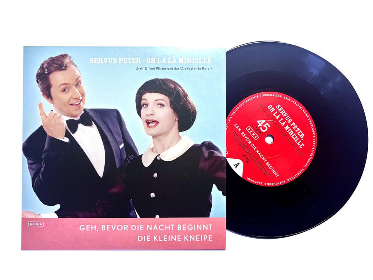 Das Bild zeigt eine Schallplatte mit dem Titel "Servus Peter - Oh la la Mireille". Die Platte ist schwarz und hat in der Mitte ein rotes Etikett, auf dem der Titel und weitere Informationen aufgedruckt sind. Neben der Platte befindet sich das Cover, das ein grafisches Design mit Farben in Blau und Rosa zeigt.  Auf dem Cover sind zwei Personen abgebildet: ein Mann und eine Frau. Der Mann trägt einen schwarzen Anzug mit einer weißen Hemd und einer schwarzen Fliege. Er hat kurzes, dunkles Haar und lächelt. Die Frau befindet sich neben ihm, trägt ein schwarzes Outfit mit einem runden Kragen und hat lockige, dunkle Haare. Beide posieren freundlich und scheinen sich in einem fröhlichen Moment zu befinden.  Unten auf dem Cover steht in weißer Schrift: "Geh, bevor die Nacht beginnt, die kleine Kneipe". Das Design vermittelt einen künstlerischen und nostalgischen Eindruck, der an die Welt der Operette und des Schlagers erinnert.