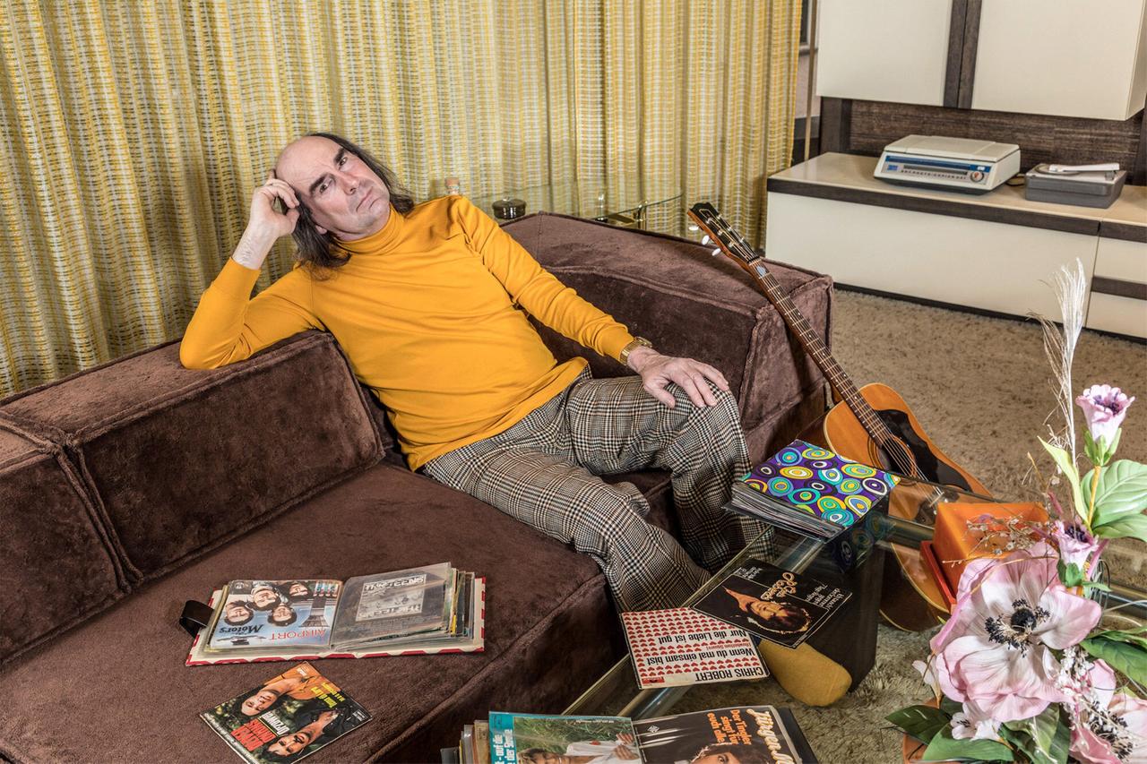 Das Bild zeigt einen Mann, der auf einem braunen Sofa sitzt. Er trägt einen leuchtend gelben Rollkragenpullover und karierte Hosen. Sein Gesichtsausdruck wirkt nachdenklich, und er hat die Hand an den Kopf gelegt. Neben ihm auf dem Sofa liegen verschiedene Schallplatten und Magazine, die teilweise aufgeschlagen sind. Am Rand des Sofas ist eine Gitarre zu sehen. Gegenüber dem Sofa befindet sich eine Wand mit einem scheinbar retro-inspirierten, hellen Hintergrund. In der Nähe gibt es einen Glastisch mit einer Blumenarrangement und weiteren Schallplatten.  Die Bildkomposition vermittelt ein Gefühl von Nostalgie und einer bestimmten Ästhetik aus einer früheren Zeit, wahrscheinlich den 1960er oder 1970er Jahren. Der Raum hat eine warme, aber auch leicht melancholische Stimmung.