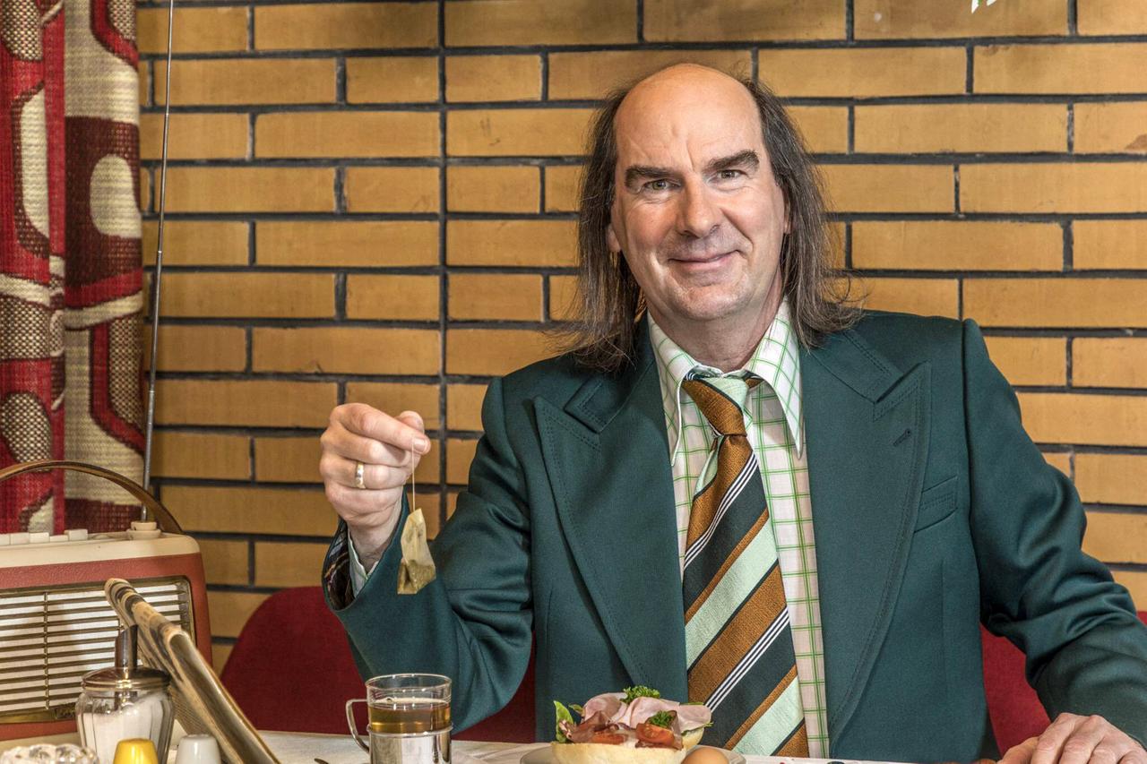 Das Bild zeigt einen Mann, der in einem Restaurant oder Café sitzt. Er hat lange, glatte, braune Haare und trägt einen auffälligen grünen Anzug mit einem karierten Hemd und einer gestreiften Krawatte in Erdtönen. In seiner rechten Hand hält er ein Teebeutel, das er gerade aus einer Tasse zieht. Vor ihm auf dem Tisch steht ein Glas mit einem Getränk, eine Tasse Tee sowie ein Teller mit einem Brötchen, das mit Wurst, Tomaten und etwas Salat belegt ist. Im Hintergrund ist eine prägnante, gemusterte Wand zu sehen, die eine retro-inspirierte Atmosphäre schafft. Auf dem Tisch steht außerdem ein älteres Radio. Die gesamte Szenerie vermittelt ein nostalgisches Gefühl.