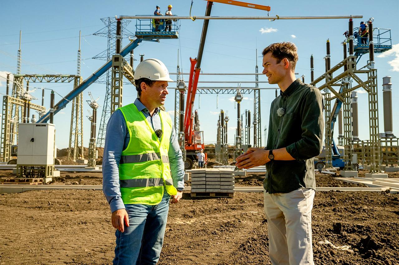 Ein Mann mit Helm und Sicherheitsweste steht mit einem anderen Mann vor einem Solarpark, der gerade im Bau ist.