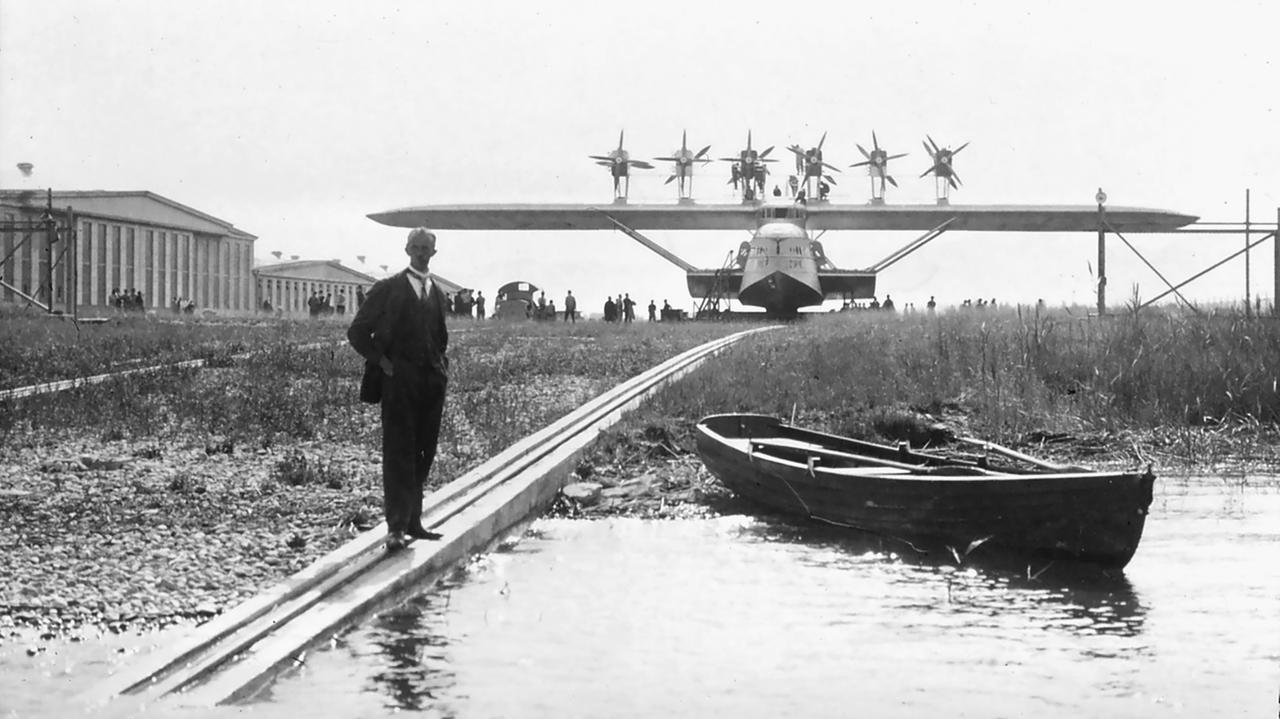 Das Bild zeigt einen historischen Flugplatz in Altenrhein, Schweiz, umgeben von einer leicht hügeligen Landschaft und Wasser. Im Vordergrund steht ein Mann, der elegant gekleidet ist, mit einer Hand in der Hosentasche. Er beobachtet ein großes Flugboot, das auf Schienen zu sehen ist und von mehreren Propellern an den Tragflächen angetrieben wird. Das Flugzeug hat eine elegante Form und ist deutlich in der Mitte des Bildes positioniert.  Links und im Hintergrund sind mehrere Gebäude zu erkennen, die eine industrielle Architektur aufweisen, vermutlich Hangars oder Werkstätten. Eine kleine Holzboot liegt am Ufer des Gewässers, das den Bereich durchzieht. Gras und kleinere Pflanzen wachsen rund um die Schienen, die das Flugboot stützen.  Im Hintergrund sind zusätzliche Personen zu sehen, die sich um das Flugzeug versammeln. Die gesamte Szene scheint in einer klaren, sonnigen Umgebung aufgenommen worden zu sein, was durch die helle, aber leicht verschwommene Bildqualität suggeriert wird.