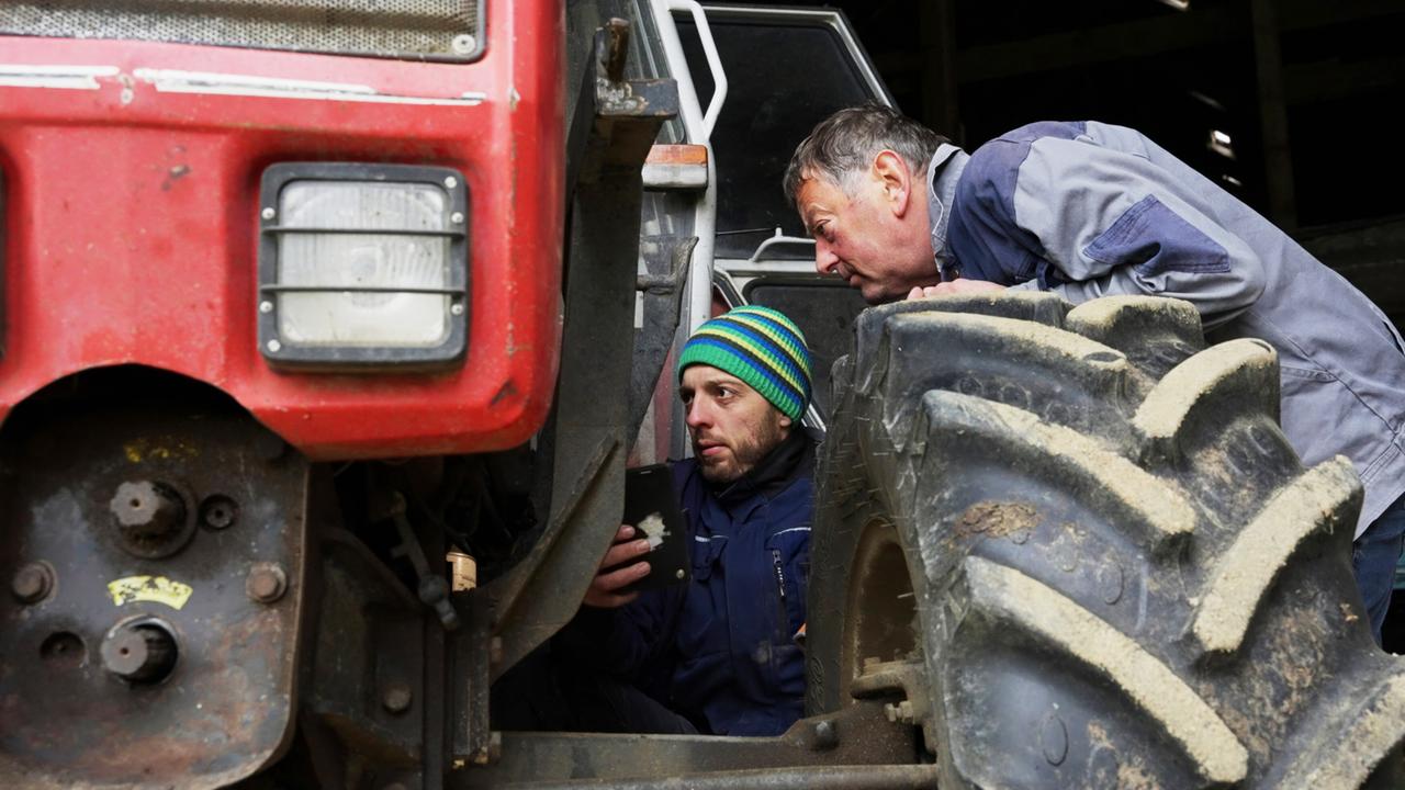 Das Bild zeigt zwei Männer, die sich unter einem roten Traktor befinden. Der Traktor hat große, dreidimensionale Reifen mit sichtbaren Rillen und Schmutz. Der Mann auf der linken Seite sitzt halb unter dem Traktor, hält ein Gerät in der Hand und trägt eine blaue Arbeitsjacke sowie eine Mütze mit blauen und grünen Streifen. Der andere Mann, der rechts über ihn gebeugt ist, hat kurze graue Haare, trägt eine graue Arbeitsjacke und schaut auf den anderen Mann und das Gerät. Im Hintergrund ist der Traktor weiter sichtbar, mit einem Teil des Kabinensystems, das teilweise im Schatten steht. Das Licht scheint natürlich und die Umgebung wirkt ländlich und landwirtschaftlich geprägt.