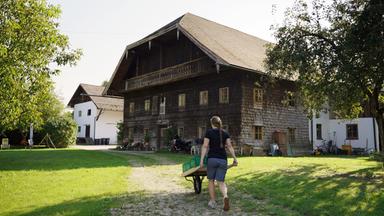 Das Bild zeigt einen traditionellen Bauernhof in Lengau, Innviertel, Österreich. Im Vordergrund ist eine Person zu sehen, die einen Wagen mit einer Holzplatte und grünen Kisten schiebt. Die Person trägt ein schwarzes T-Shirt und kurze Hosen, hat lange, geflochtene Haare und läuft auf einem gepflasterten Weg, der durch eine grüne Wiese führt.  Im Hintergrund sind zwei Gebäude sichtbar: das größere, ältere Haus ist mit dunklen Holzschindeln verkleidet und hat eine große Dachüberstände, während das kleinere, hellere Gebäude rechteckig ist und mit weißen Wänden gestaltet ist. Zwischen den Gebäuden gibt es Grünflächen und einige Pflanzen sowie Geräte, die zur Landwirtschaft genutzt werden. Die gesamte Szenerie vermittelt eine ruhige, ländliche Atmosphäre an einem sonnigen Tag.