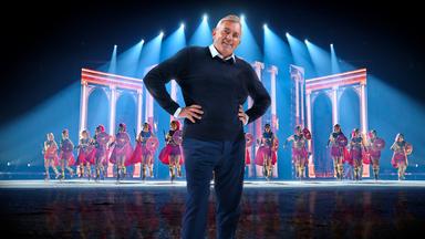 Das Bild zeigt eine Szene, die wie eine Bühnenperformance aussieht. Im Vordergrund steht ein Mann in einem dunkelblauen Pullover und einer blauen Hose. Er hat graue Haare und lächelt in die Kamera, wobei er seine Hände in die Hüften gestemmt hat. Hinter ihm sind eine Gruppe von Einkunstläufer:innen in Kostümen zu sehen, die wie römische Soldaten gekleidet sind. Sie tragen rote Umhänge, goldene Brustpanzer und Helme sowie rote Schilde, dazu Schlittschuhe. Der Hintergrund der Bühne hat eine Kulisse, die an römische Architektur erinnert, mit hohen Säulen und Bögen. Über ihnen sind blaue Lichter zu sehen, die auf die Bühne gerichtet sind und eine dramatische Beleuchtung erzeugen.