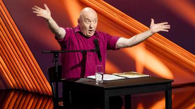 Horst Evers sitzt auf der linken Bildseite an einem Tisch. Er hebt beide Arme an der Seite. Der Hintergrund ist rot beleuchtet.  