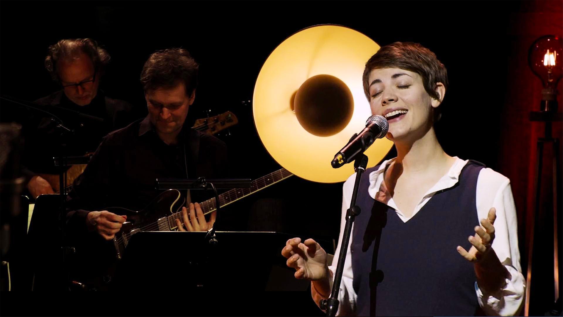 Die Sängerin FEE. steht singend und lächelnd auf der Bühne. Hinter ihr zwei Gitarristen.