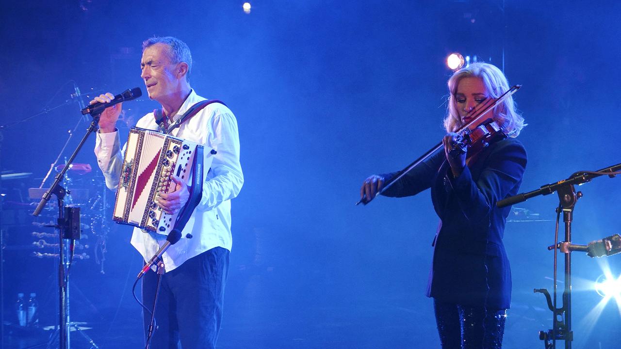 Auf dem Bild sind zwei Musiker auf einer Bühne während eines Konzerts im Salzburger Festspielhaus zu sehen. Der Mann steht links und hält ein Akkordeon in der linken Hand und ein Mikrofon in der rechten Hand. Er trägt ein helles Hemd und hat ein konzentriertes Gesicht, während er singt. Die Frau rechts spielt Violine und hat lange blonde Haare. Sie trägt ein schwarzes Outfit. Im Hintergrund sind Lichter und unscharfe Silhouetten von weiteren Musikern oder Instrumenten zu erkennen. Die gesamte Szenerie ist von einem blauen Lichtschein umgeben, der eine stimmungsvolle Atmosphäre erzeugt.