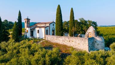 Luftaufnahme der Cappella San Gaetano, die von hohen Zypressen und einer historischen Steinmauer umgeben ist, eingebettet in eine grüne Hügellandschaft.