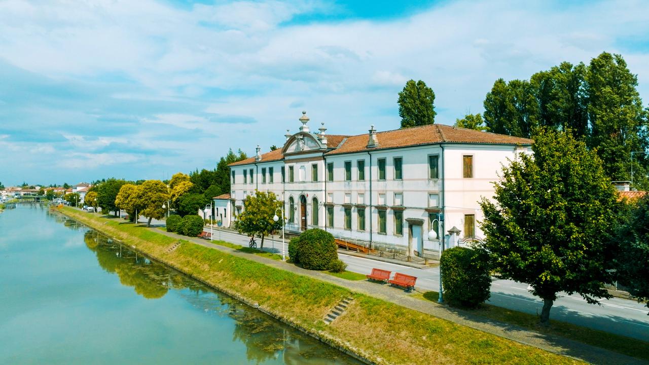 Villa an der Brenta, gelegen an einem ruhigen Kanal, mit Bäumen und einer Promenade entlang des Ufers.