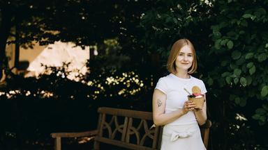 Vanessa steht vor einer grünen Hecke und einer Parkbank. Sie hat ein Eis in der Hand und schaut lächelnd in die Kamera.
