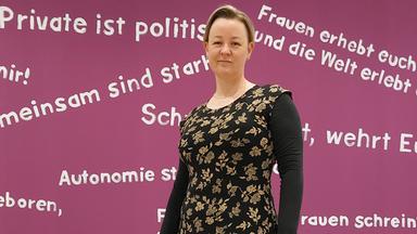 Sandra Schmitz vor beerenfarbener Wand mit diversen feministischen Schriftzügen.