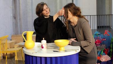 Cara Hoffmann und Pfarrerin Rebekka Wackler stehen hinter einem Taufbecken. Pfarrerin Rebekka Wackler trägt Talar und hat die rechte Hand erhoben, um den Taufsegen durchzuführen. Cara Hoffmann hat sich nach vorn gebeugt, um den Segen zu empfangen.