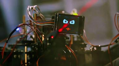 Ein spinnenartiger kleiner Roboter mit vielen Kabeln und Drähten sitzt vor einer Keyboard-Tastatur und drückt mit einem seiner Greifarme auf eine Taste. Als Kopf hat er einen kleinen Bildschirm, auf dem sein lachendes Gesicht zu sehen ist. Der Hintergrund ist unscharf, auf einem Bildschirm ist eine bunte Computeranimation zu erkennen.