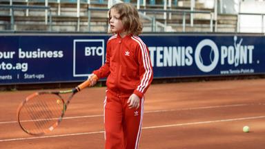 Ein Kind im roten Trainingsanzug auf dem Tennisplatz