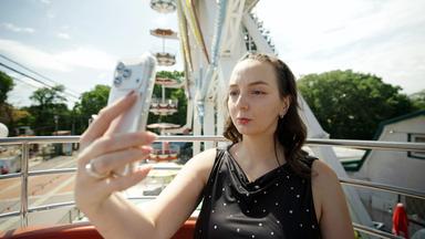 Das Bild zeigt eine junge Frau, die in ein Smartphone schaut, während sie anscheinend ein Selfie von sich macht. Sie sitzt in einer Fahrgeschäfte, möglicherweise einem Riesenrad, da im Hintergrund Teile des Rades sowie Sitzplätze zu sehen sind. Die Frau trägt ein schwarzes Oberteil mit kleinen, glänzenden Herzen. Ihr Haar ist lockig und fällt locker über ihre Schultern. Die Umgebung ist hell erleuchtet, und es ist ein klarer, sonniger Tag. Hinter ihr ist das grüne Laub von Bäumen sichtbar sowie bunte Fahrgeschäfte und Tische, die darauf hindeuten, dass es sich um einen Freizeitpark oder ein Jahrmarkt handelt.