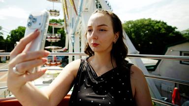 Das Bild zeigt eine junge Frau, die in ein Smartphone schaut, während sie anscheinend ein Selfie von sich macht. Sie sitzt in einer Fahrgeschäfte, möglicherweise einem Riesenrad, da im Hintergrund Teile des Rades sowie Sitzplätze zu sehen sind. Die Frau trägt ein schwarzes Oberteil mit kleinen, glänzenden Herzen. Ihr Haar ist lockig und fällt locker über ihre Schultern. Die Umgebung ist hell erleuchtet, und es ist ein klarer, sonniger Tag. Hinter ihr ist das grüne Laub von Bäumen sichtbar sowie bunte Fahrgeschäfte und Tische, die darauf hindeuten, dass es sich um einen Freizeitpark oder ein Jahrmarkt handelt.