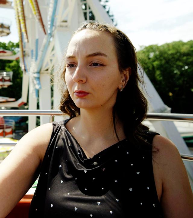 Das Bild zeigt eine junge Frau, die in ein Smartphone schaut, während sie anscheinend ein Selfie von sich macht. Sie sitzt in einer Fahrgeschäfte, möglicherweise einem Riesenrad, da im Hintergrund Teile des Rades sowie Sitzplätze zu sehen sind. Die Frau trägt ein schwarzes Oberteil mit kleinen, glänzenden Herzen. Ihr Haar ist lockig und fällt locker über ihre Schultern. Die Umgebung ist hell erleuchtet, und es ist ein klarer, sonniger Tag. Hinter ihr ist das grüne Laub von Bäumen sichtbar sowie bunte Fahrgeschäfte und Tische, die darauf hindeuten, dass es sich um einen Freizeitpark oder ein Jahrmarkt handelt.