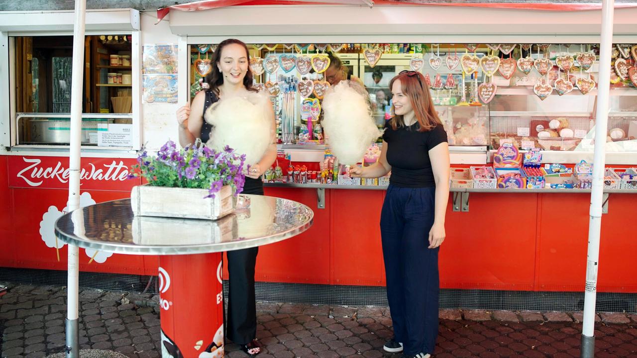 Das Bild zeigt zwei Frauen, die in einem Freizeitpark vor einem Stand aus Zuckerwatte stehen. Die Standfläche ist rot und trägt die Aufschrift "Zuckerwatte". Hinter den Frauen sind verschiedene Süßwaren und Snacks sichtbar, die in Regalen und auf Theken platziert sind.   Links von den Frauen hält eine Person in einem schwarzen, ärmellosen Oberteil eine große Zuckerwatte in der Hand. Sie hat lange, glatte Haare und lächelt. Die andere Person, die rechts steht, trägt ein schwarzes T-Shirt und dunkelblaue, weite Hosen. Sie hält ebenfalls eine Zuckerwatte und blickt lächelnd zur Seite. Der Tisch vor ihnen ist rund und hat eine glänzende Oberfläche.  Im Hintergrund sind aufgehängte Lebkuchenherzen und andere Süßigkeiten zu erkennen. Der Bereich wirkt lebhaft und festlich, passend zu einem Tag im Freizeitpark.