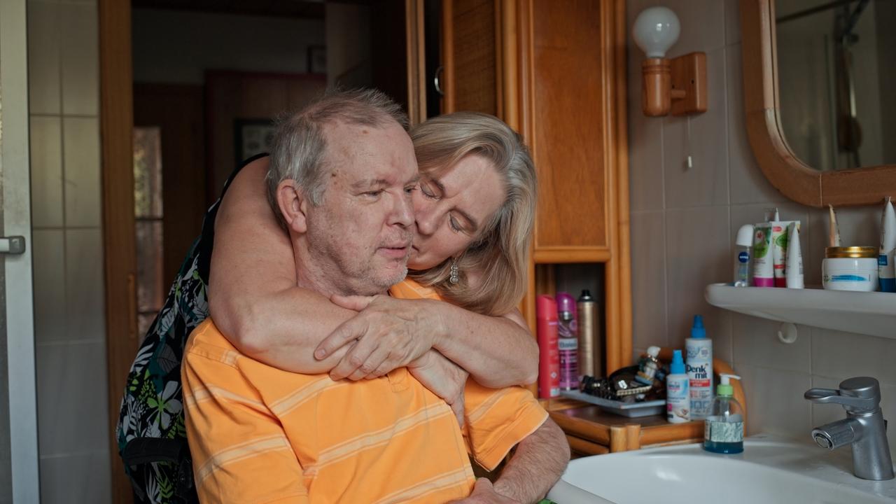 Ein still vor sich hin schauender Mann in gelbem Poloshirt sitzt im Rollstuhl vor dem Waschbacken in einem engen Badezimmer. Eine Frau beugt sich von hintern über ihn und umarmt ihn.