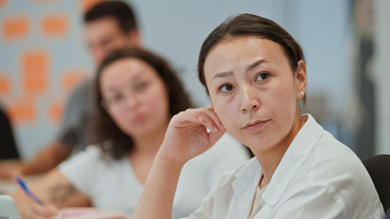 An einem Tisch sitzen zwei junge Frauen und ein junger Mann unterschiedlicher Herkunft, die konzentriert auf jemandem außerhalb des Bildes schauen. Die der Kamera zunächst sitzende Frau ist scharf, die anderen beiden etwas unscharf.
