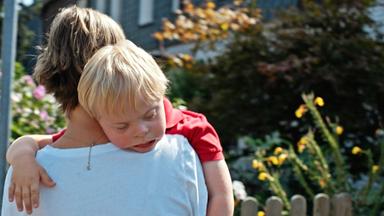 Rückansicht einer Frau in weißem T-Shirt, die einen kleinen schlafenden Jungen über die Schulter trägt. Im Hintergrund sieht man blühende Blumen in Vorgärten von Häusern.