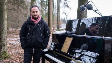 Ein Mann in Winterjacke steht neben einem Klavier im Wald.
