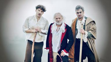 Szene aus "Das Wesen des Lebens" von Iida Turpeinen: Drei Männer stehen nebeneinander. Zwei von ihnen stützen sich auf dickeren Stöcken ab. Der Mann in der Mitte ist älter und hat weiße Haare und einen weißen Vollbart. 