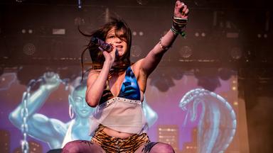 Eine leicht bekleidete Frau mit Tattoos auf den Beinen hockt auf einer Bühne uns singt in ein Mikro, während sie die linke Faust nach oben reckt.