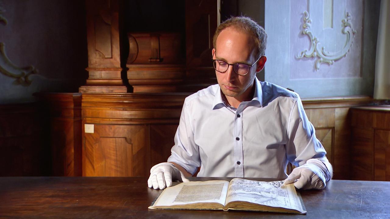 Ein Mann mit Brille und weißen Handschuhen betrachtet ein altes Buch mit Illustrationen an einem Holztisch in einer historischen Bibliothek.