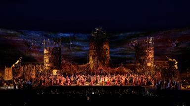 Totale auf die Bühne von “Il Trovatore” in der Arena di Verona. Im Vordergund sieht man das Orchester, auf der Bühne stehen drei große Türme mit Fahnen darauf. Darunter sind viele Menschen auf der Bühne versammelt.