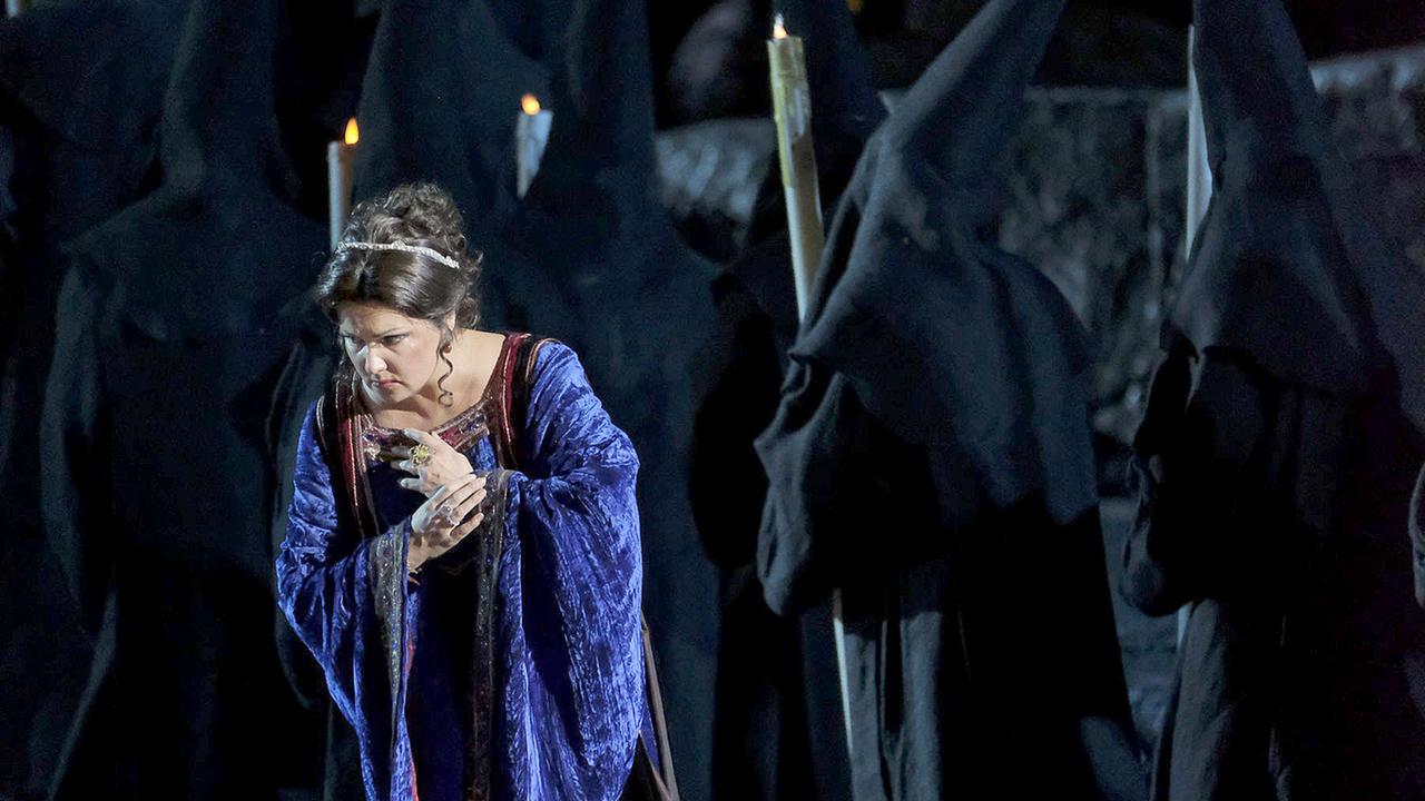 Blick auf die Bühne von “Il Trovatore” in der Arena di Verona. Anna Netrebko steht in gebeugter Haltung im Vordergrund. Sie trägt ein weinrotes Kleid. Im Hintergrund laufen Nonnen mit brennenden Kerzen. 