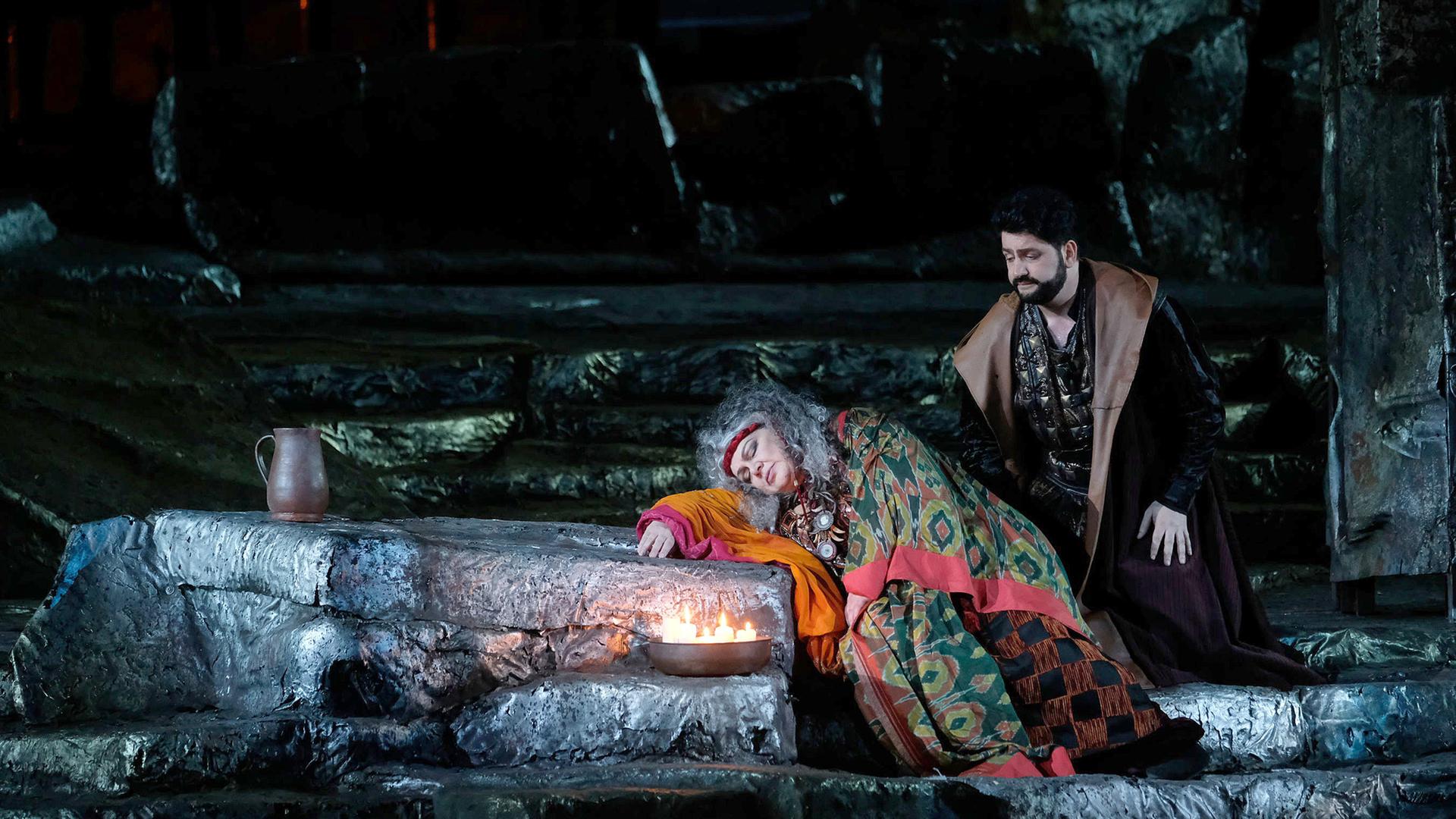 Blick auf die Bühne von “Il Trovatore” in der Arena di Verona. Dolora Zajick lehnt mit geschlossenen Augen auf einem Felsen. Sie ist ein buntes Tuch gehüllt. Rechts hinter ihr kniet Yusif Eyvazov und blickt besorgt auf sie.