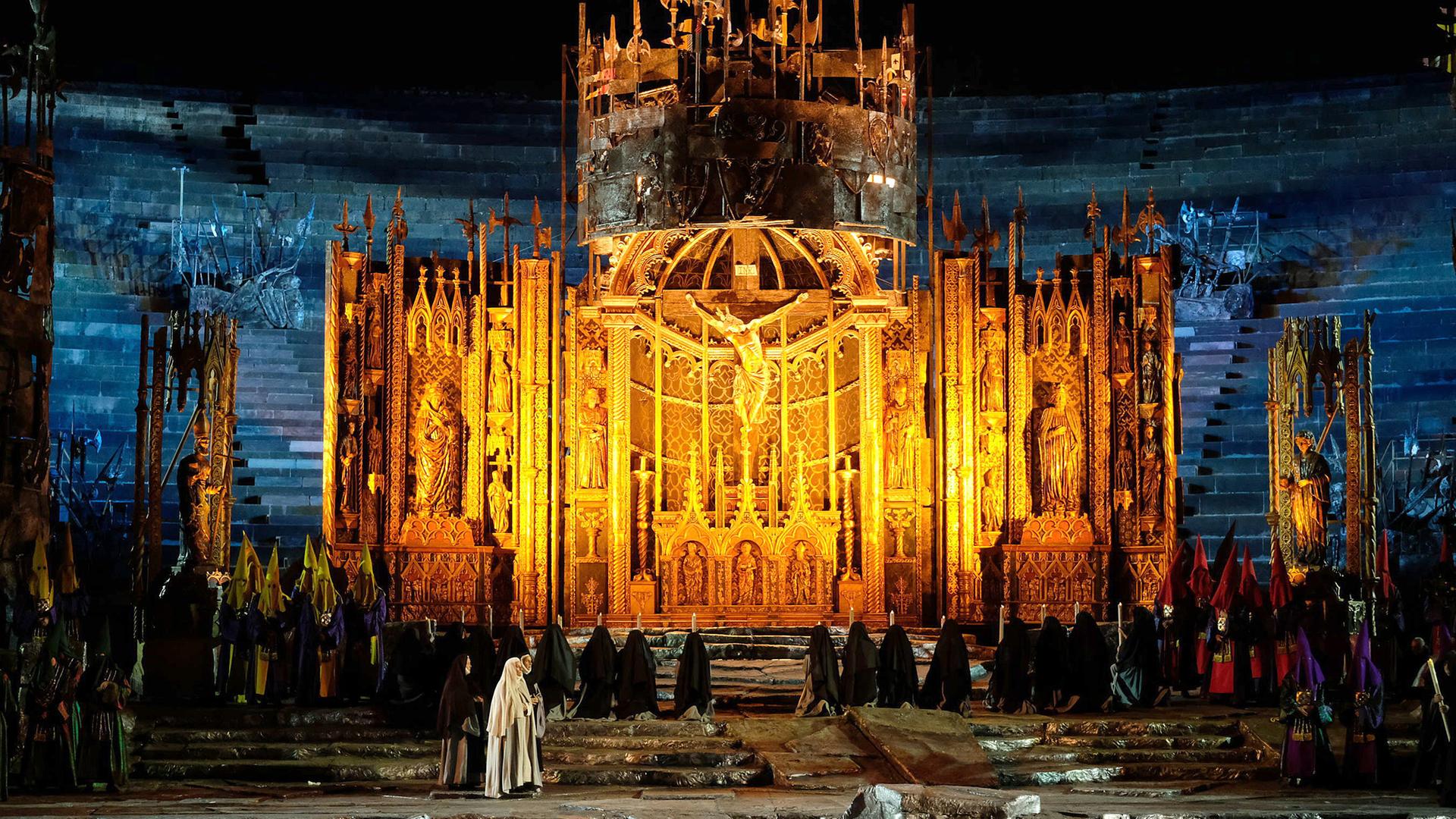 Blick auf die Bühne von “Il Trovatore” in der Arena di Verona. Im Hintergrund steht ein großer goldener Altar. Vor dem Altar steht eine große Gruppe an Nonnen mit Kerzen in der Hand. Im Vordergrund stehen drei Nonnen. Die mittlere Nonne trägt ein weißes Gewand. 