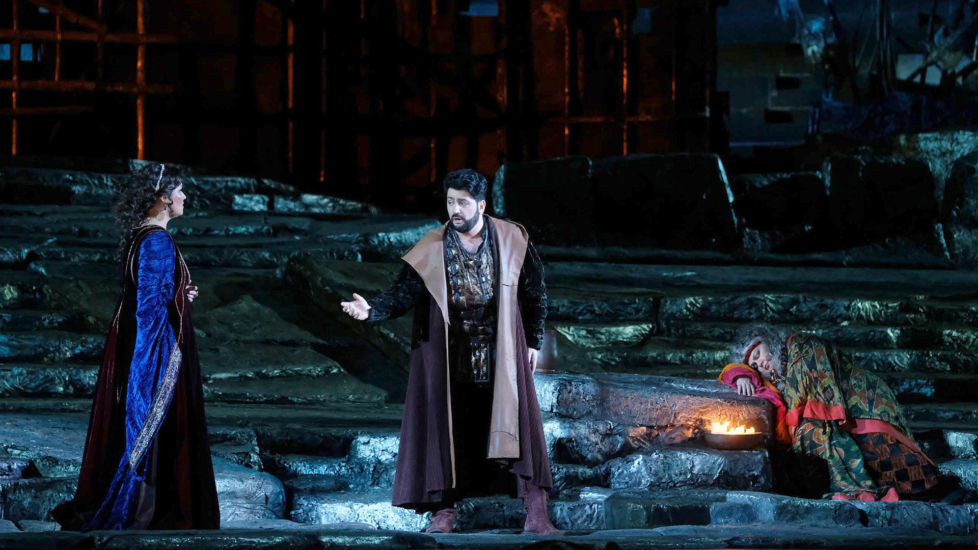 Blick auf die Bühne von “Il Trovatore” in der Arena di Verona. Im Vordergrund stehen Anna Netrebko (li) und Yusif Eyvazov (mi). Sie trägt ein lila-weinrotes Kleid und ist von der Seite zu sehen. Jusiy Eyvazov ist von vorne zu sehen und hebt seine rechte Hand in Richtung Anna Netrebko. Auf der linken Seite der Bühne liegt Dolora Zajick an einem Felsvorsprung gelehnt. Sie ist ein buntes Tuch gehüllt.   