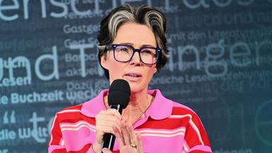 Eine Frau mittleren Alters mit kurzen dunklen Haaren, die vorne graue Strähnen haben, großer blauer Kunststoffbrille und pink gestreiftem Pullover, spricht in ein Mikrofon.