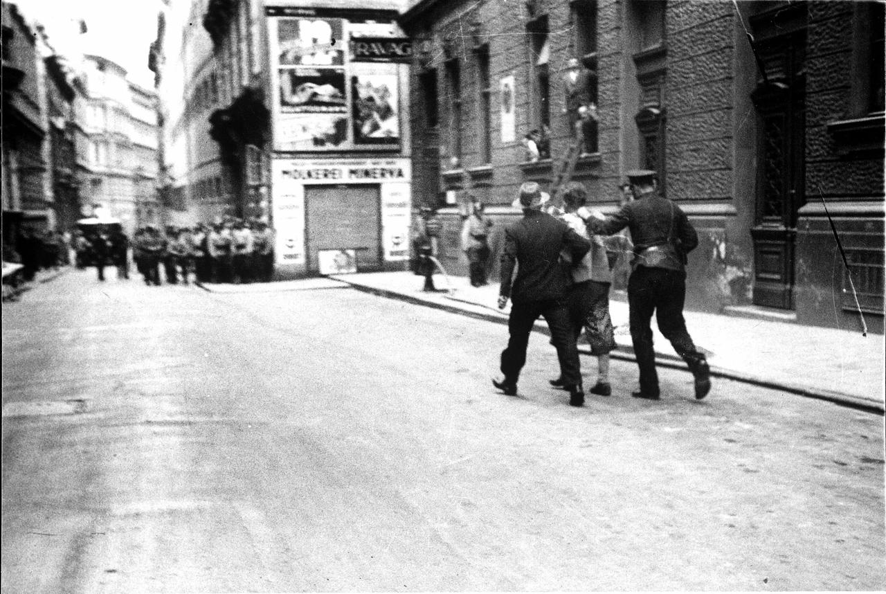 Das Bild zeigt eine historische Szene aus dem Juli-Putsch 1934 in Österreich. Im Vordergrund sind zwei Männer zu sehen, die einen festgenommene Person, einen Putschisten, begleiten. Der festgenommene Mann trägt eine hellfarbene Hose und ein Hemd. Einer der beiden Begleiter ist in einer uniformähnlichen Kleidung, der andere trägt Anzug und Hut.  Im Hintergrund erkennt man eine Straßenansicht, die von Gebäuden gesäumt ist, deren Fassaden zum Teil mit Plakaten bedeckt sind. Auf der linken Seite sind einige Soldaten oder Polizisten in Uniform aufgereiht, die die Situation beobachten. Die Straßen sind leer, und es gibt Anzeichen von Unruhe in der Umgebung.  Die Bildqualität ist schwarz-weiß und hat einen historischen Charakter, was die Atmosphäre der damaligen Zeit widerspiegelt.