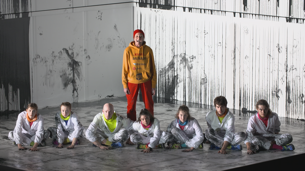 Theaterszene mit Aktivist*innen auf der Bühne. Im Vordergrund sitzen in weißer Schutzkleidung 7 junge Männer und Frauen im Schneidersitz mit festgeklebten Händen auf der Bühne. Hinter ihnen steht eine Frau mit roter Mütze und orangenen Kapuzenpulli und schreit. Auf dem Pulli sind die Logos von DHL, Amazon, Hermes, Ups u.a. abgedruckt.