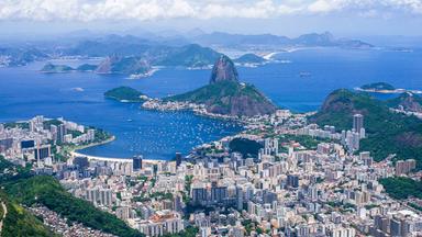 Luftaufnahme von Rio de Janeiro mit dicht bebauten Stadtvierteln, der Guanabara-Bucht und dem markanten Zuckerhut zwischen grünen Bergen und blauem Meer.