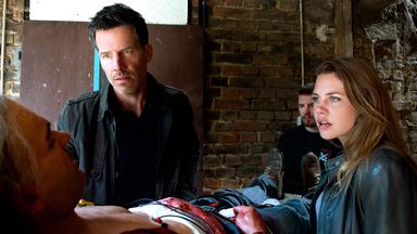 "Im Wald (2) - Ein Taunuskrimi": Oliver von Bodenstein (Tim Bergmann) und Pia Sander (Felicitas Woll) stehen an einer Liege, auf der ein verletzter Mann zu sehen ist. Beide schauen den Verletzten fassungslos an.