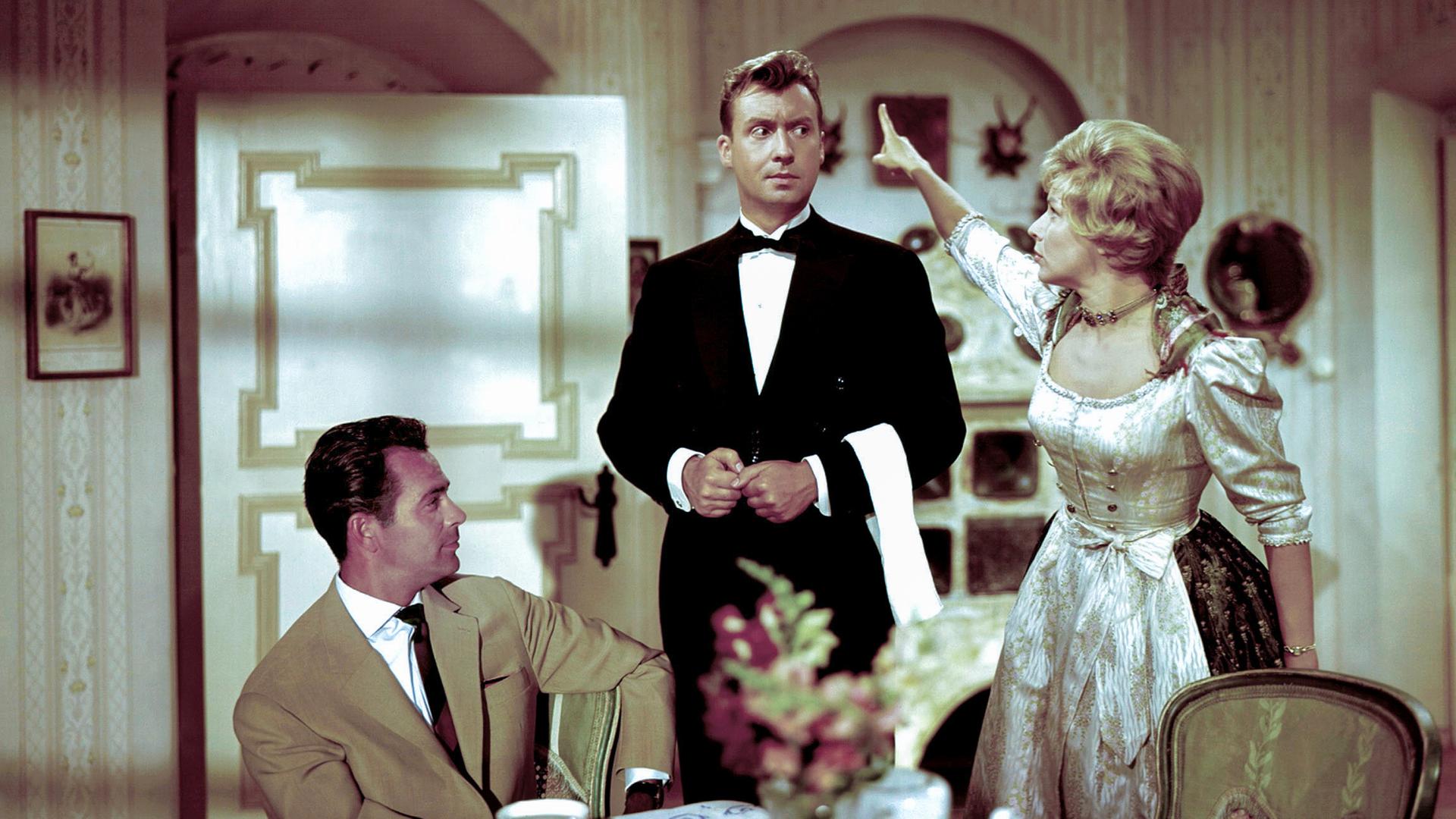 Ein Mann sitzt an einem Tisch, neben ihm ein Kellner, der von einer Frau mit einer Zeigegeste aus dem Raum geschickt wird.